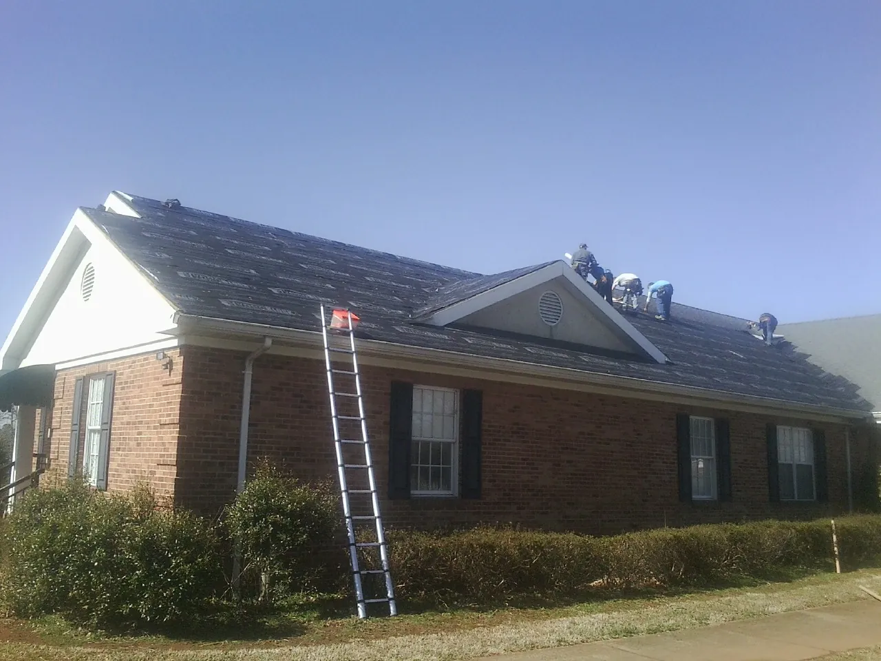 Crew tearing shingles off a brick ranch roof with a ladder set — Emergency Roof Repair work by our crew in Lorane