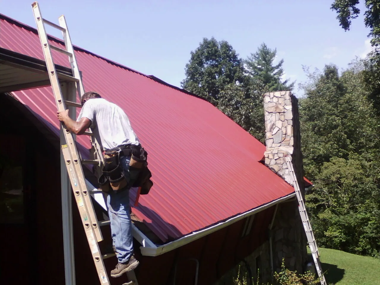 Roofer on a ladder beside a red metal roof and stone chimney — Soffit & Fascia Repair by Roofing Brothers in Lorane